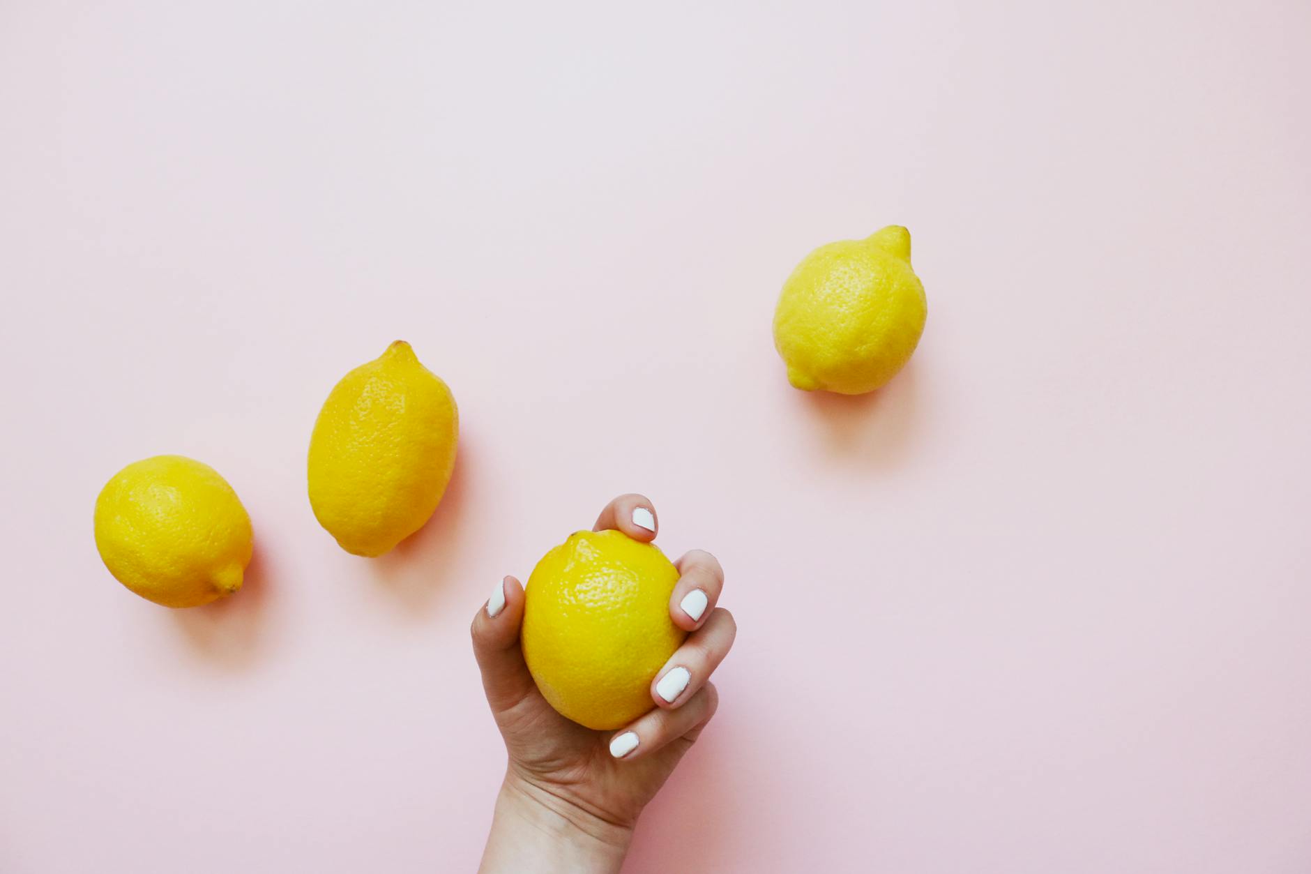 Una mano con unghie bianche che tiene un limone su uno sfondo rosa morbido, circondato da tre limoni aggiuntivi.