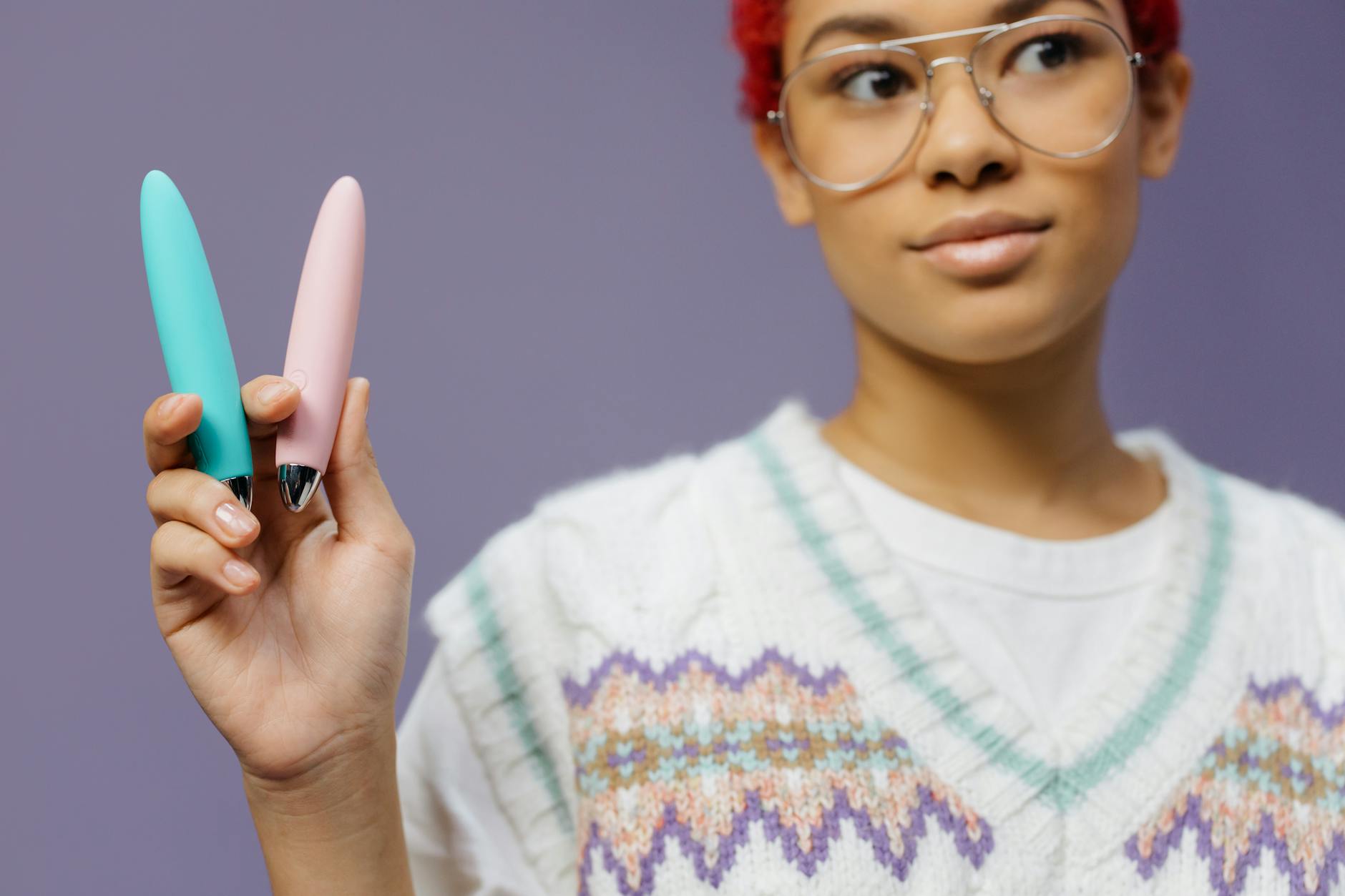 Una giovane donna con occhiali tiene due vibratori colorati su uno sfondo viola