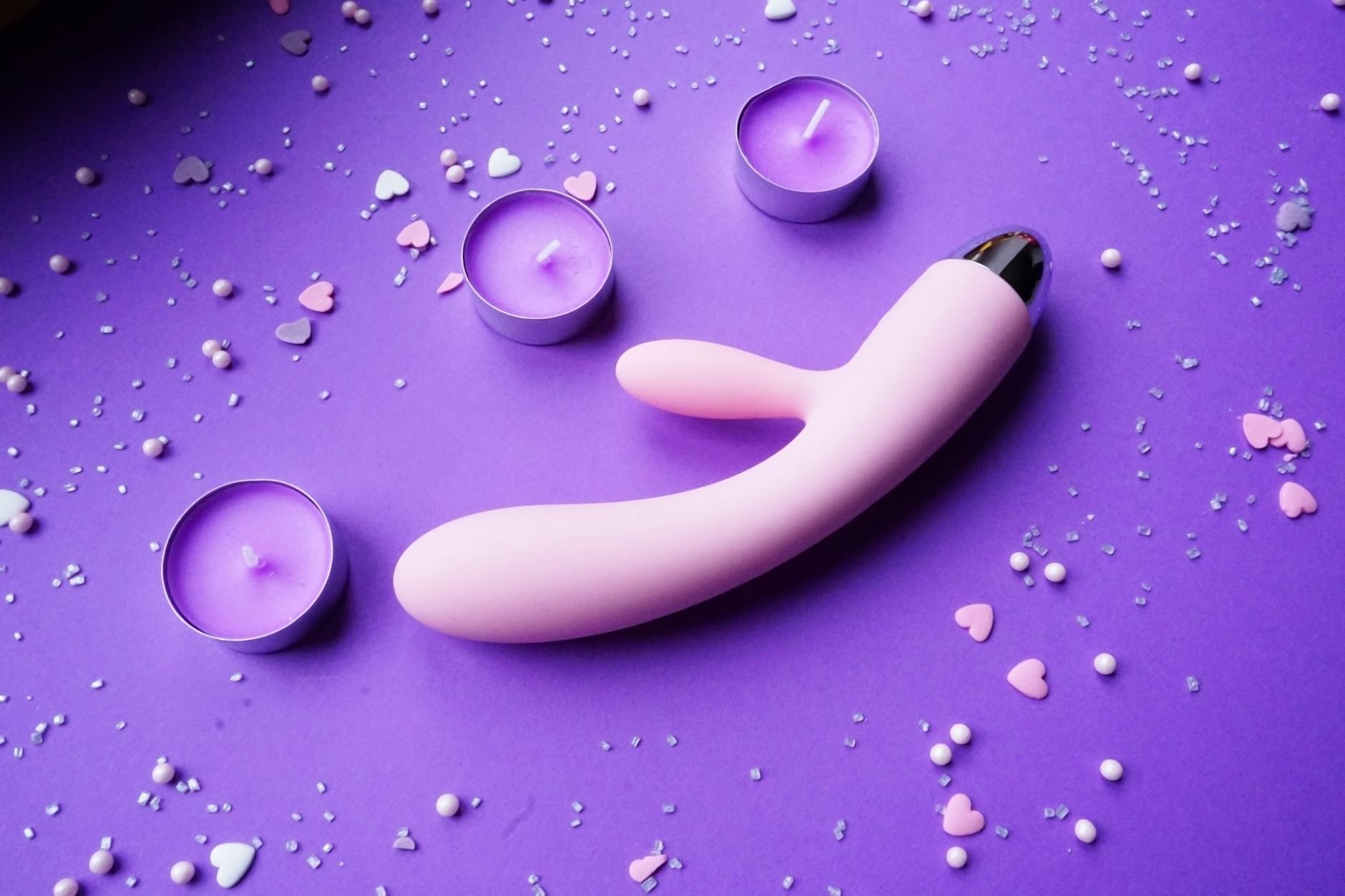 Vibratore rosa su sfondo viola con coriandoli a forma di cuore e candele per un'atmosfera romantica.